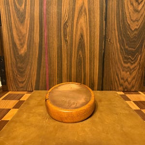 May include: A small, round wooden container with a light brown finish. The container has a flat, circular top and a curved, rounded body. The background features a wooden surface with a checkerboard pattern and a vertical wooden panel.