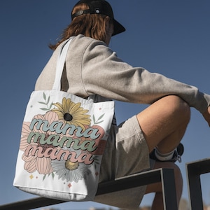 May include: White tote bag with a floral design and the word "mama" in a retro font. The flowers are in shades of yellow, pink, and blue. The bag has a simple design and is carried over the shoulder.