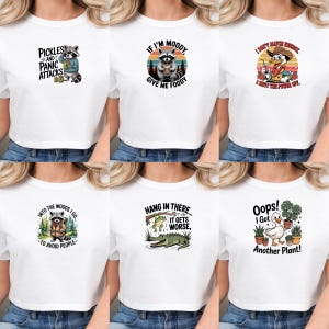 May include: A collection of six white t-shirts featuring various graphic prints: a raccoon with pickles, a raccoon with food, a duck in a cowboy hat, a raccoon in the woods, a frog and alligator, and a duck holding a potted plant with text.