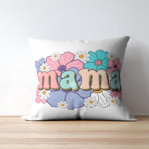 May include: White square throw pillow with the word "mama" in a retro font, surrounded by a floral design. The flowers are in shades of pink, blue, purple, and white, creating a colorful and cheerful aesthetic. The pillow is on a wooden surface.