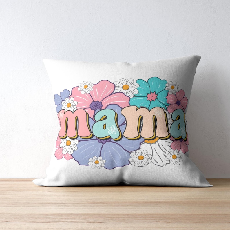 May include: White square throw pillow with the word "mama" in a retro font, surrounded by a floral design. The flowers are in shades of pink, blue, purple, and white, creating a colorful and cheerful aesthetic. The pillow is on a wooden surface.