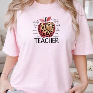 May include: A light pink t-shirt featuring a graphic of an apple with a leopard print pattern and a gold glitter heart in the center. The apple is surrounded by the words blessed, love, protect, comfort, care, kind, inspire, teach, guide, and support.