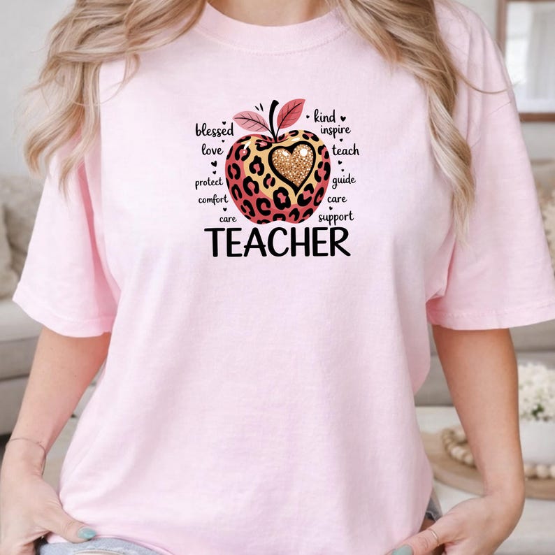 May include: A light pink t-shirt featuring a graphic of an apple with a leopard print pattern and a gold glitter heart in the center. The apple is surrounded by the words blessed, love, protect, comfort, care, kind, inspire, teach, guide, and support.