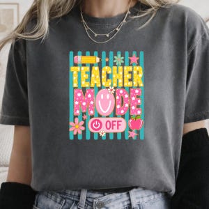 May include: A charcoal gray t-shirt featuring a graphic print with vertical teal stripes. The text reads TEACHER MODE OFF in yellow and pink. The design includes a yellow pencil, a pink smiley face, flowers, stars, and a red apple icon.