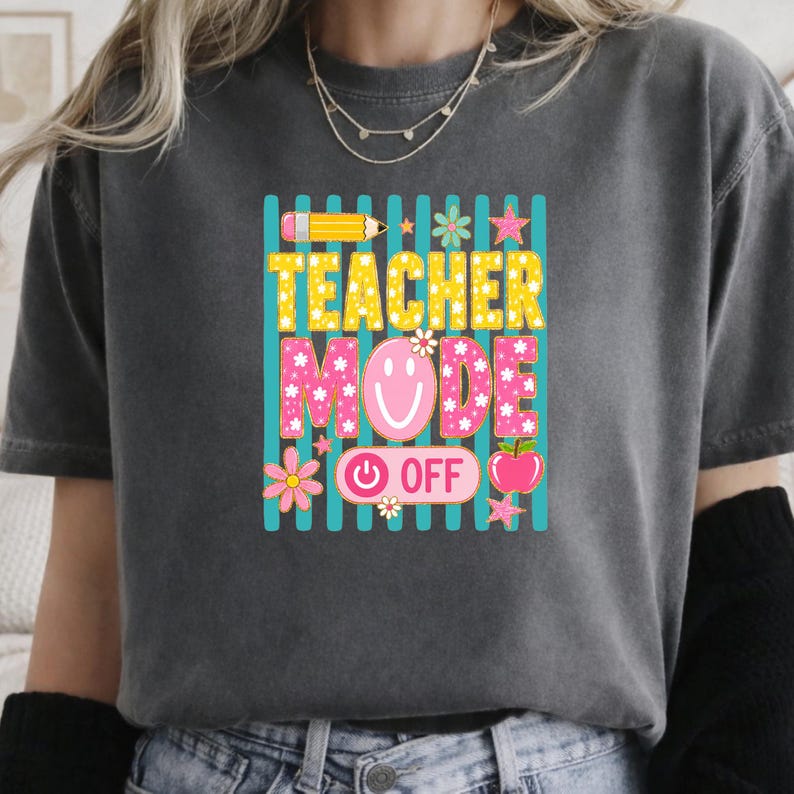 May include: A charcoal gray t-shirt featuring a graphic print with vertical teal stripes. The text reads TEACHER MODE OFF in yellow and pink. The design includes a yellow pencil, a pink smiley face, flowers, stars, and a red apple icon.