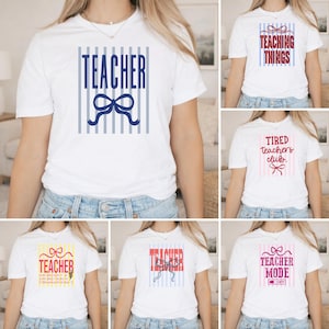 May include: A white short-sleeve t-shirt featuring various teacher-themed graphic designs. Each shirt displays vertical stripes, a bow illustration, and text such as TEACHER, TEACHING THINGS, TIRED TEACHERS CLUB, or TEACHER MODE in red, blue, or pink.