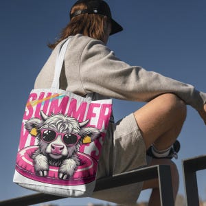 Op de afbeelding: Een witte draagtas met een cartoon koe met een zonnebril en een roze zwemband. Het woord "SUMMER" is in het roze gedrukt met een regenboogboog erboven. De tas heeft witte bandjes.