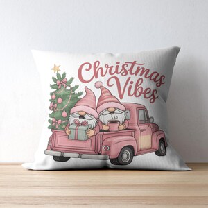 May include: White pillow with a Christmas theme. Features a pink truck carrying two gnomes, a Christmas tree, and the words "Christmas Vibes" in pink script. The gnomes wear pink hats and hold a gift and a mug.