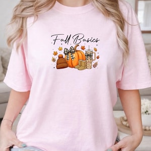 K&ouml;nnte beinhalten: Ein hellrosa T-Shirt mit dem Text Fall Basics in schwarzer Schreibschrift. Die Grafik zeigt einen orangefarbenen K&uuml;rbis mit einer schwarz-wei&szlig; karierten Schleife, eine braune Strickm&uuml;tze, beige Stiefel und einen Becher mit Leopardenmuster, umgeben von Herbstbl&auml;ttern.