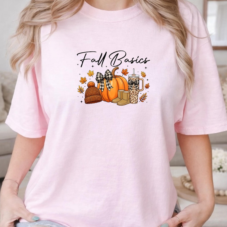 K&ouml;nnte beinhalten: Ein hellrosa T-Shirt mit dem Text Fall Basics in schwarzer Schreibschrift. Die Grafik zeigt einen orangefarbenen K&uuml;rbis mit einer schwarz-wei&szlig; karierten Schleife, eine braune Strickm&uuml;tze, beige Stiefel und einen Becher mit Leopardenmuster, umgeben von Herbstbl&auml;ttern.