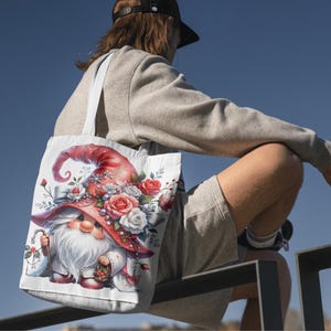 May include: White tote bag featuring a whimsical gnome design. The gnome wears a red hat adorned with roses and carries a cane. The artwork includes a white beard, a floral arrangement, and a charming, detailed illustration.