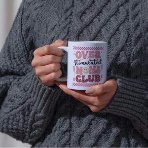 May include: A white ceramic mug with the text "OVER STIMULATED MOMS CLUB" in pink and red lettering. The mug features a smiley face and a checkered pattern. The mug is held by a person wearing a gray sweater.