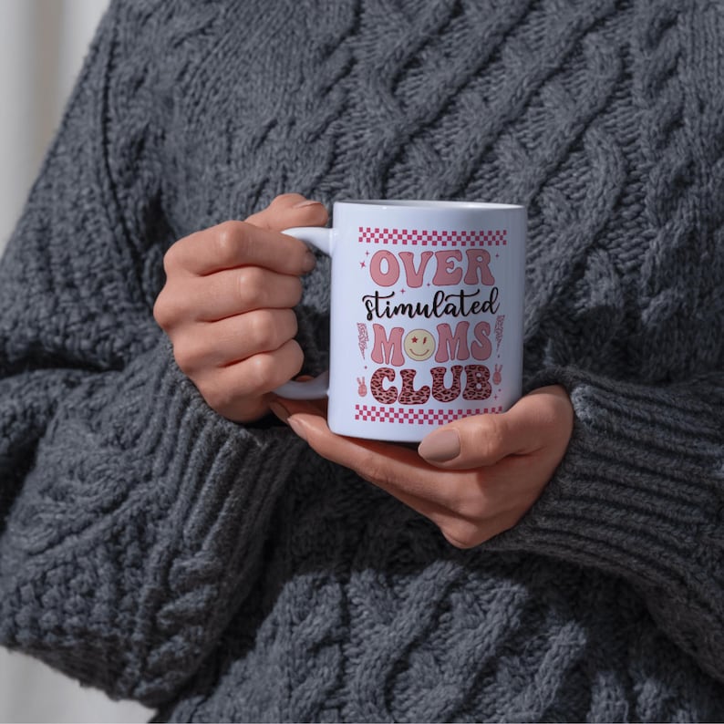 May include: A white ceramic mug with the text "OVER STIMULATED MOMS CLUB" in pink and red lettering. The mug features a smiley face and a checkered pattern. The mug is held by a person wearing a gray sweater.