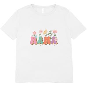 May include: White t-shirt featuring the word "MAMA" in colorful, retro bubble letters, with flowers sprouting from the letters. The design includes pink, teal, orange, and purple hues, with a brown outline.