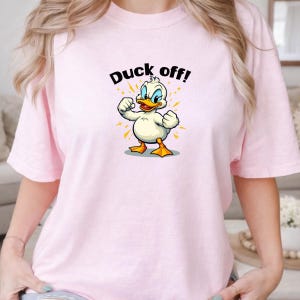 May include: A light pink t-shirt featuring a graphic of a white duck with orange feet and a beak, standing with its fists clenched. Above the duck, the text "Duck off!" is printed in black letters, surrounded by small yellow star-like accents.
