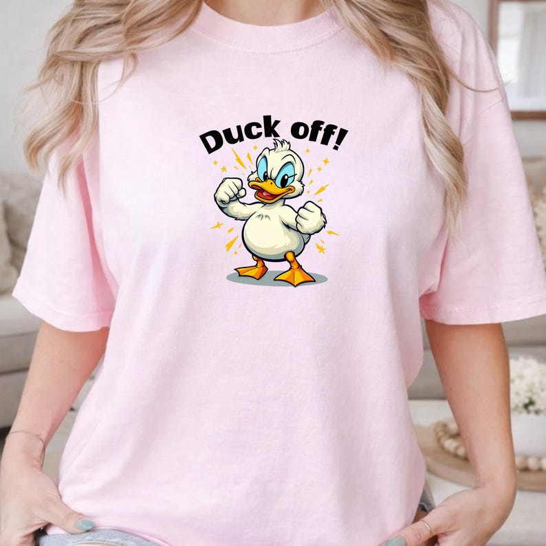 May include: A light pink t-shirt featuring a graphic of a white duck with orange feet and a beak, standing with its fists clenched. Above the duck, the text "Duck off!" is printed in black letters, surrounded by small yellow star-like accents.