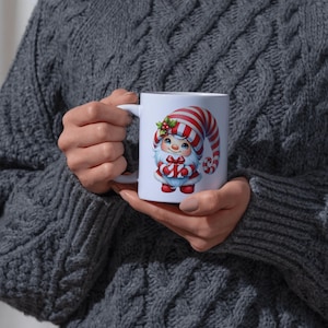 May include: White ceramic mug featuring a whimsical Christmas gnome design. The gnome wears a red and white striped hat and holds a wrapped gift. The mug has a white handle and is held by a person wearing a gray sweater.