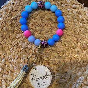 Può includere: Un braccialetto portachiavi con perline con un mix di perline blu, rosa e leopardate. Il portachiavi ha una chiusura color oro, un nappa e un ciondolo rotondo in legno con il testo "Proverbi 3:5".