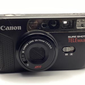 May include: A black Canon Sure Shot Tele Max 35mm film camera featuring a Canon Lens 38/70mm 1:3.5/6.0. The front of the camera displays the Canon logo, the model name in white and red text, an autofocus lens, a built-in flash, and a sliding switch.