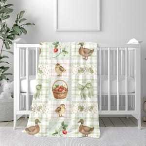 Puede incluir: Una manta para bebé con un diseño de patchwork con patos, patitos, fresas y lazos. La manta tiene un patrón de cuadros verdes y blancos con ilustraciones de acuarela. La manta está sobre una cuna blanca.