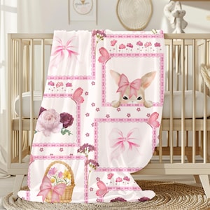 May include: A white baby blanket with a pink and floral design. The blanket features a repeating pattern of pink bows, butterflies, flowers, and a bunny with a pink bow. The blanket is draped over a wooden crib.