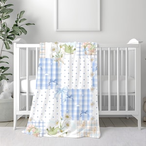 May include: A baby blanket with a patchwork design featuring blue gingham, white polka dots, and floral patterns. The blanket has light blue bows and illustrations of birds and flowers. It is draped over a white crib.