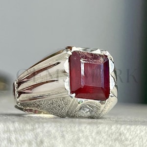 May include: A silver ring with a large, rectangular, deep red gemstone. The ring has a classic design with a textured band and a setting that securely holds the gemstone. The gemstone's facets reflect light, creating a brilliant display.