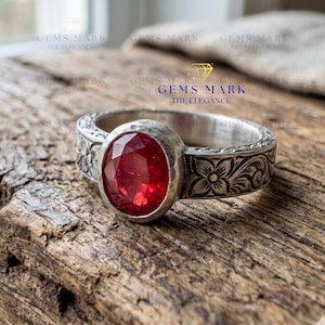 Könnte beinhalten: Silberring mit einem ovalen, facettierten roten Edelstein. Das Band weist ein Blumenmuster auf. Der Ring wird auf einer Holzoberfläche präsentiert. Der Text "GEMS MARK THE ELEGANCE" ist im Hintergrund sichtbar.