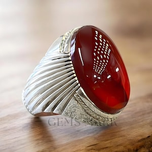 Puede incluir: Anillo de plata con una gran gema roja ovalada pulida. El anillo tiene un diseño texturizado y acanalado en los laterales y un engaste detallado alrededor de la piedra. La palabra "GEMS" es visible en el anillo.