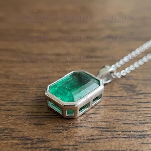 Pode incluir: Um colar com pingente de prata com uma pedra preciosa verde retangular com corte esmeralda. A esmeralda é engastada em uma moldura de prata com uma corrente delicada. O pingente é exibido em uma superfície de madeira.