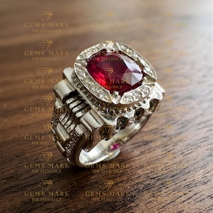 May include: A silver ring with a large, oval-cut ruby gemstone at the center, surrounded by smaller, clear stones. The ring has a detailed, textured band and is set against a wooden surface. The text "GEMS MARK THE ELEGANCE" is visible.