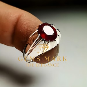 Puede incluir: Un anillo plateado con una gran gema roja oscura de corte ovalado. El anillo tiene un diseño detallado y texturizado en la banda y alrededor de la gema. El texto "GEMS MARK THE ELEGANCE" es visible.