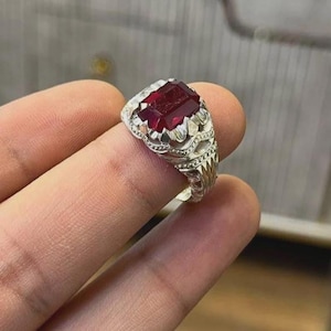 Puede incluir: Un anillo de plata con un diseño ornamentado y una gran gema rectangular de color rojo oscuro. El anillo está sostenido por un dedo, mostrando los intrincados detalles del trabajo en metal y el color vibrante de la piedra.