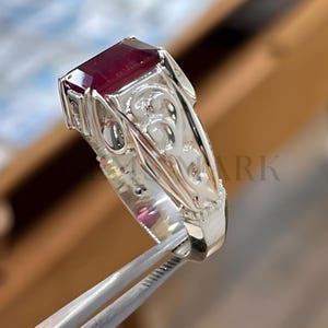 May include: A silver ring with a large, rectangular, deep red gemstone. The ring has an ornate, filigree design on the band. The gemstone is held in place by prongs. The ring is being held by a pair of tweezers.