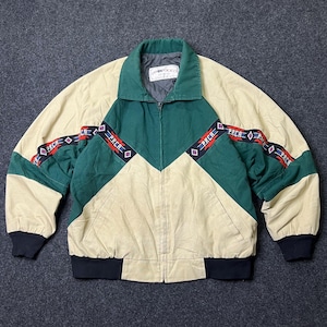 Op de afbeelding: Een vintage jas in beige, groen en zwart. De jas heeft een colorblock-ontwerp met een groene chevron op de borst en mouwen. De mouwen hebben een rood-zwarte band met het woord "PRCA" herhaald. De jas heeft een kraag en een ritssluiting.