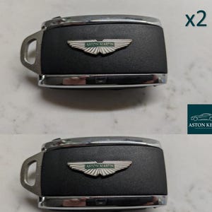 Peut inclure: Deux clés de voiture Aston Martin. Chaque clé a un corps noir avec une garniture argentée et une boucle argentée. Le logo Aston Martin est centré sur le corps noir. Le texte "x2" est dans le coin supérieur droit.