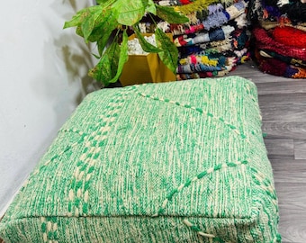 May include: A square, green woven pouf with a textured surface and white and green stitched details. The pouf is a soft, decorative floor cushion. The background includes a plant and a stack of colorful rugs.