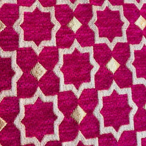 May include: Close-up of a vibrant fuchsia fabric with a repeating pattern of gold and white star-shaped designs. The texture appears plush, suggesting a decorative textile.