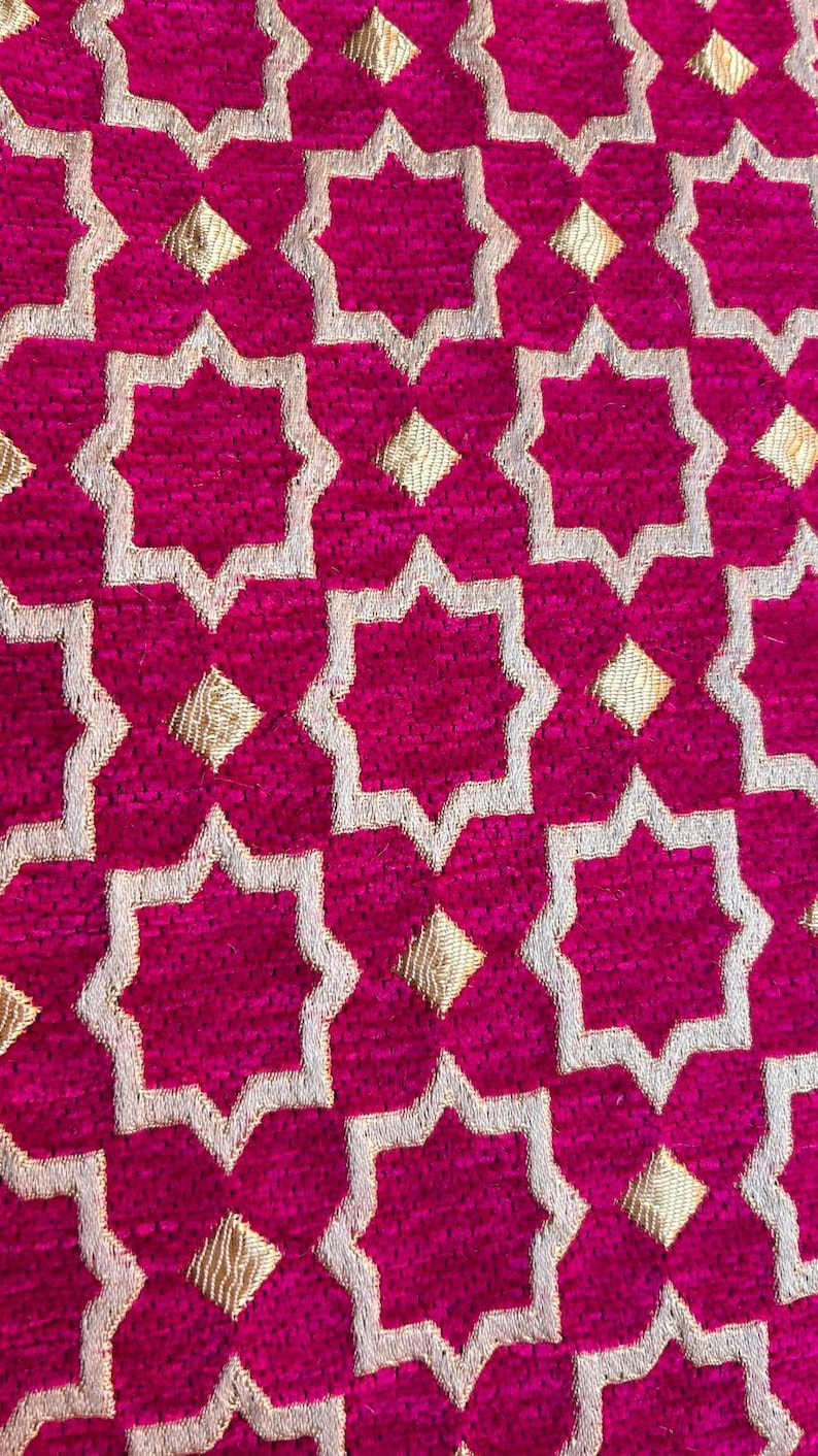 May include: Close-up of a vibrant fuchsia fabric with a repeating pattern of gold and white star-shaped designs. The texture appears plush, suggesting a decorative textile.
