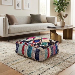 Könnte beinhalten: Ein quadratischer Bodenpouf mit einem lebendigen, mehrfarbigen Patchwork-Design auf der Oberseite und einer Mischung aus blauen, roten und türkisfarbenen Stoffstreifen an den Seiten. Die Textilkonstruktion zeigt eine gewebte Struktur mit Mustern.