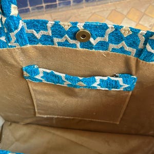 May include: A turquoise tote bag with a geometric pattern in silver and gold. The bag has a gold-colored interior and a small pocket with a matching handle. A magnetic snap closure is visible.