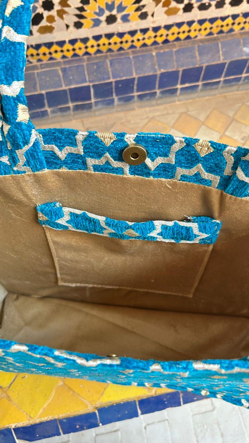 May include: A turquoise tote bag with a geometric pattern in silver and gold. The bag has a gold-colored interior and a small pocket with a matching handle. A magnetic snap closure is visible.