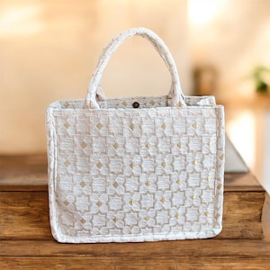 May include: Cream-colored tote bag with a geometric pattern of gold diamonds. The bag has a structured shape, a top handle, and a button closure. The fabric has a textured appearance, suggesting a woven material.