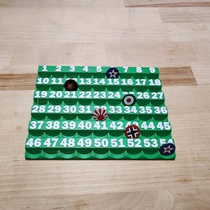 May include: A green tiered organizer with numbered slots, holding various round tokens with military insignia. The numbers 1-54 are printed on the organizer. The tokens feature symbols like a star, a bullseye, and a swastika.