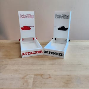 May include: Two white wooden game piece holders for the board game Axis & Allies. One has a red tank graphic and the word "ATTACKER" in red. The other has a black tank graphic and the word "DEFENDER" in black.