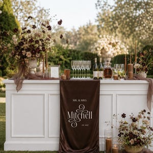 Puede incluir: Una barra blanca cubierta con un camino de mesa de terciopelo marrón oscuro con el texto impreso en blanco MR. & MRS. Mitchell est. 2027. La barra contiene copas de vino, dos botellas, un decantador y velas marrones entre flores.