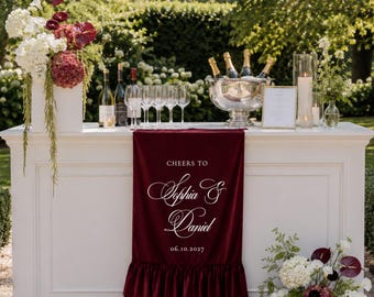 Puede incluir: Una barra blanca con un camino de mesa de terciopelo color burdeos profundo con un borde con volantes. El camino muestra el texto blanco CHEERS TO, Sophia & Daniel, y 06.10.2027. La barra está decorada con arreglos florales y cristalería.