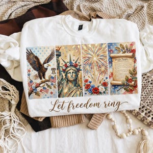Può includere: Felpa bianca con un design patriottico raffigurante un'aquila, la Statua della Libertà, fuochi d'artificio e la Dichiarazione di Indipendenza. Le parole "Let freedom ring" sono scritte in basso.