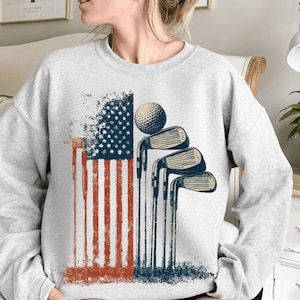 Può includere: Una felpa grigio chiaro con un design a bandiera americana invecchiata con strisce rosse e bianche e stelle blu. Mazze da golf e una pallina da golf sono posizionate a destra della bandiera, creando un tema golfistico patriottico.