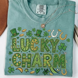 May include: A teal Comfort Colors t-shirt with a "Lucky Charm" graphic in green and yellow. The design includes shamrocks, horseshoes, coins, and top hats. A silver necklace with a small pendant rests on the shirt.